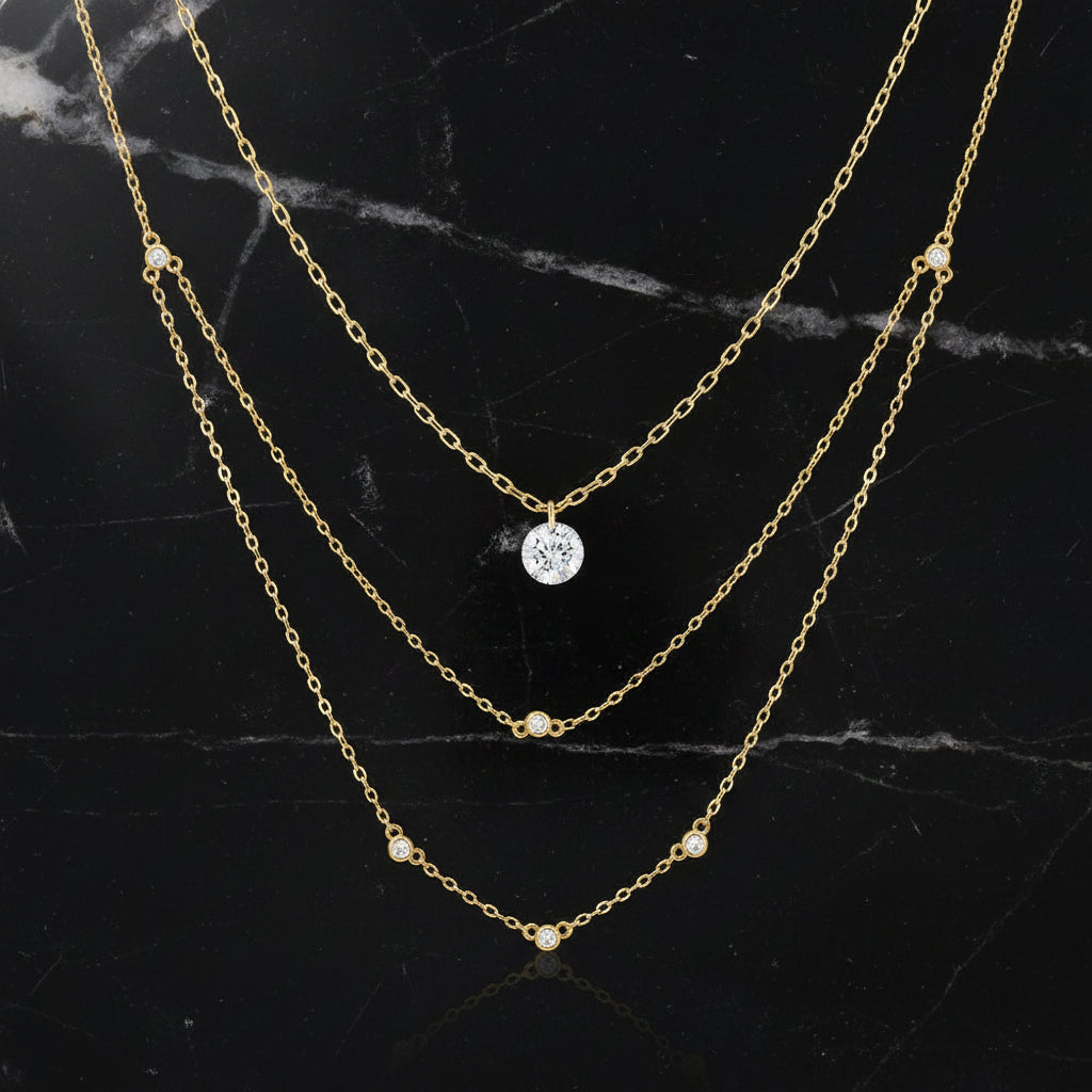 A three-layered gold necklace set by Canner, featuring a delicate chain, a mid-length chain with a single round-cut clear stone pendant, and a longer chain with small gold bead accents. The necklace is displayed on a clean white background with the Canner logo in the top-left corner and a 'Sterling Silver 925' hallmark stamp in the bottom-right.